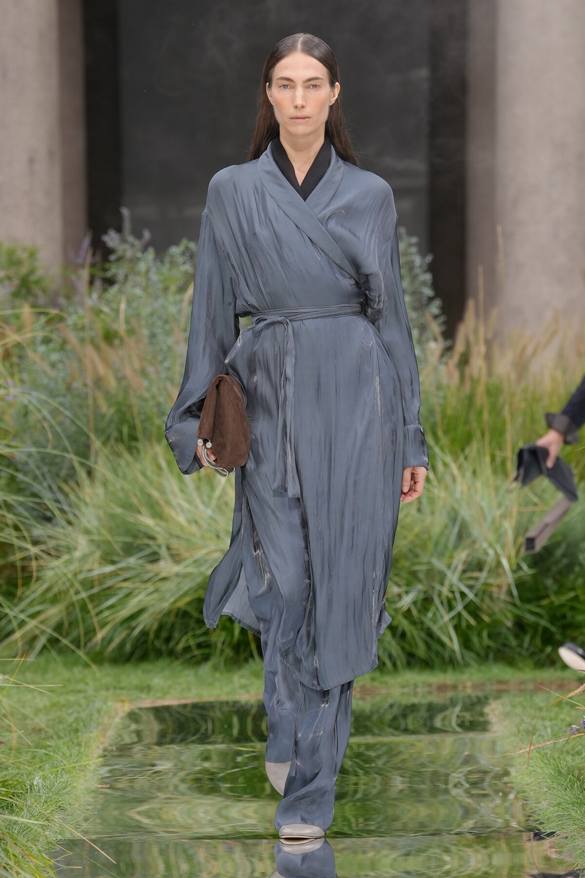 Female model walking a runway in a blue-gray wrap trench with belt, holding a brown clutch outdoors.
