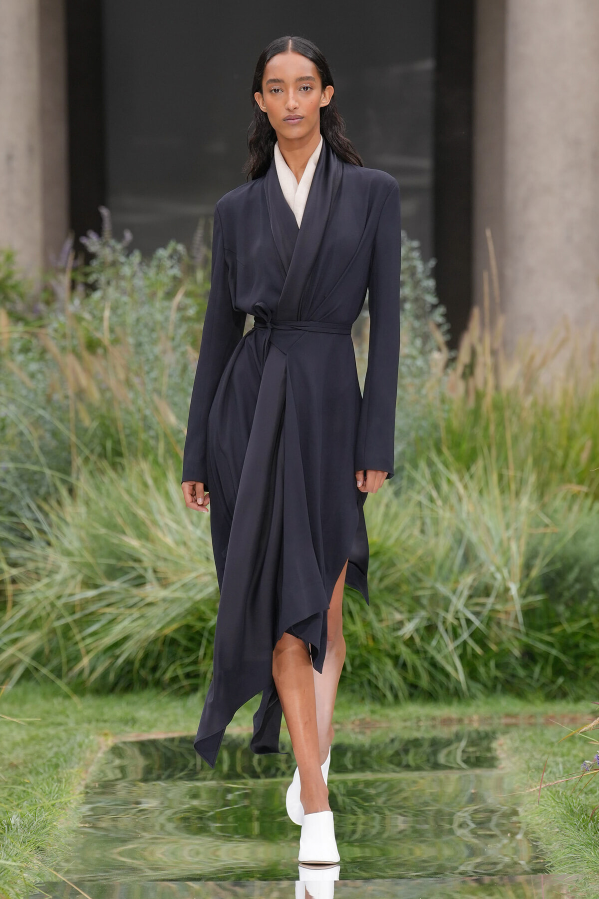 Model walks a dark navy wrap dress with a white inner collar along a reflective runways path with greenery around her.