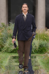 Man wearing a navy double-breasted blazer and brown trousers walks on a reflective runway outdoors, holding a hat in one hand.