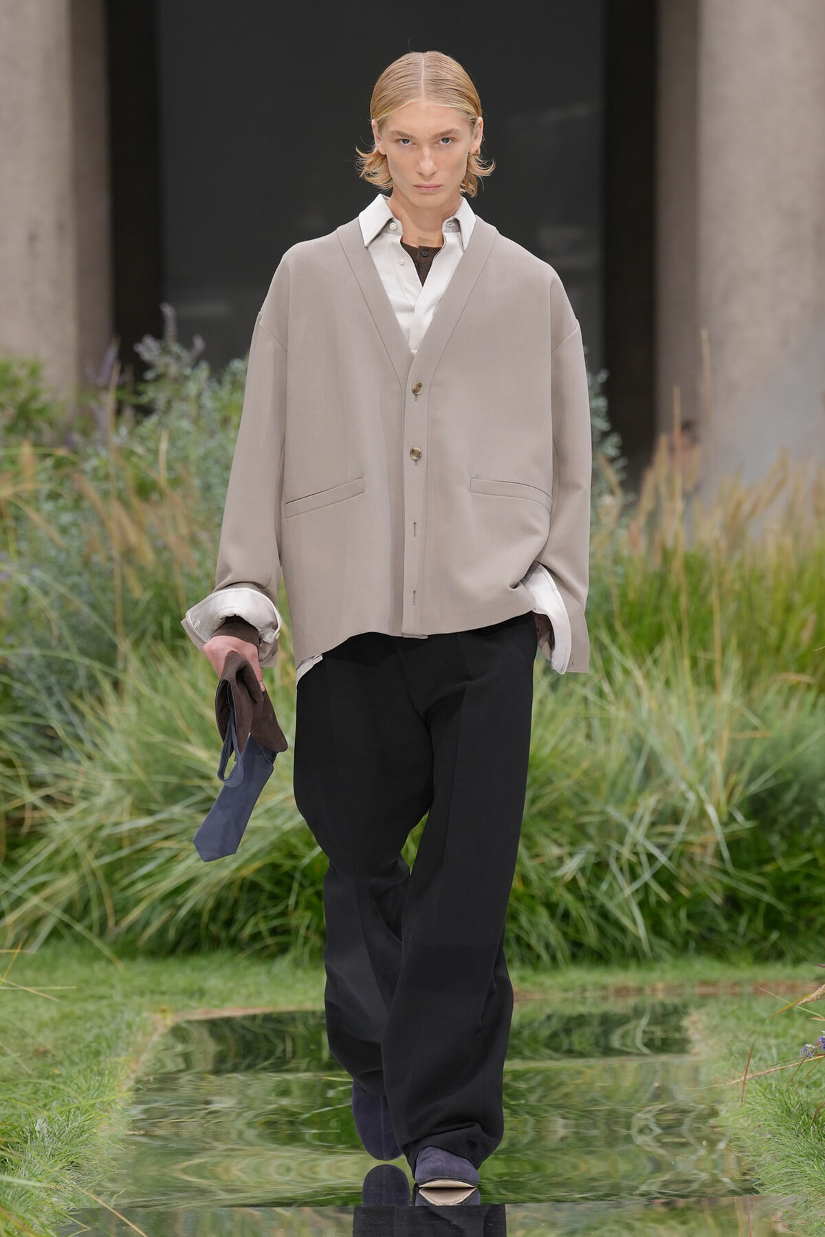 Model walking an outdoor runway in a loose beige cardigan over a white shirt and black trousers, holding a navy scarf in one hand.
