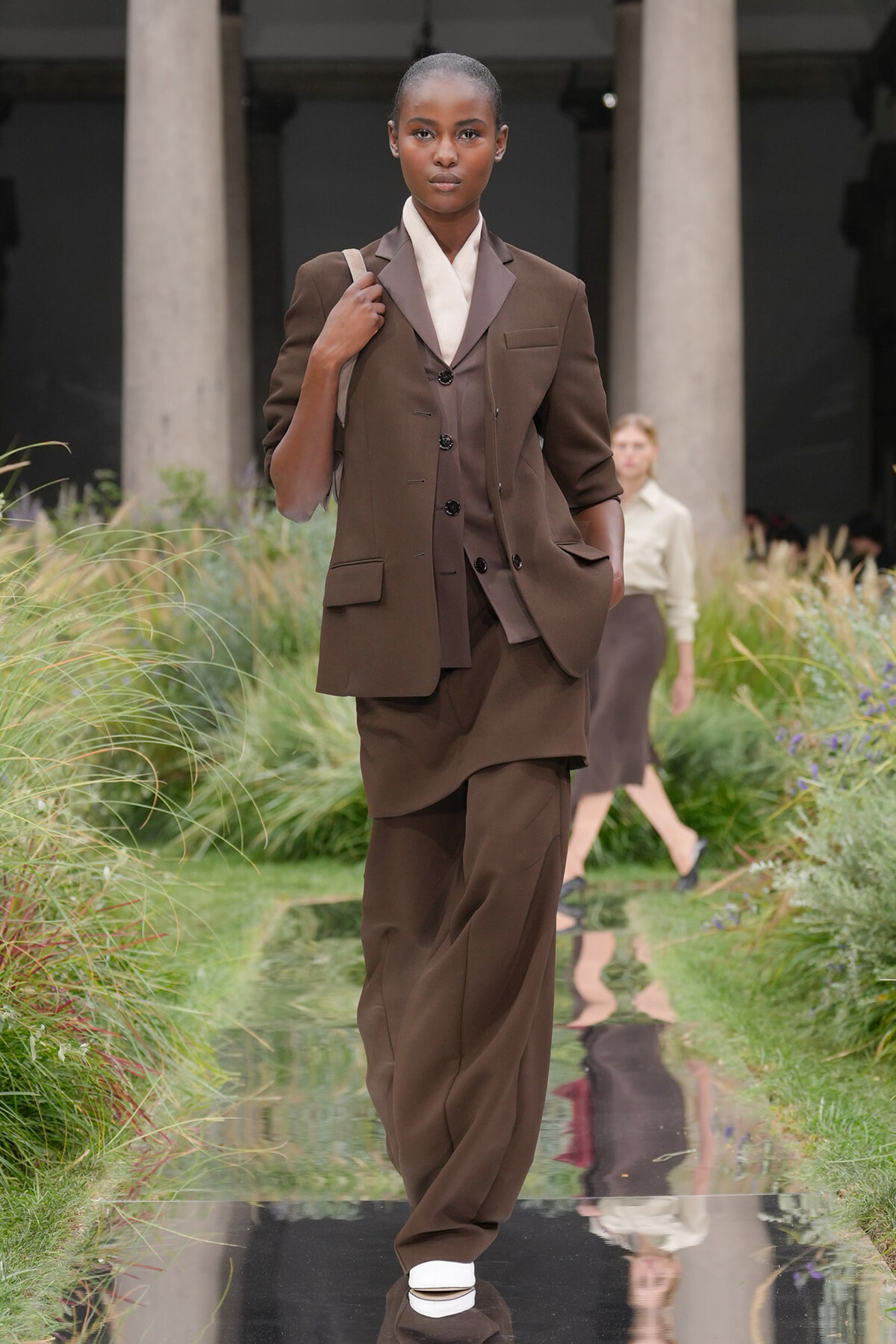 Model walking a fashion runway in a brown three-piece suit with blazer, vest, and wide trousers, carrying a beige shoulder bag.