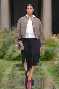 Model walking on a runway in an oversized taupe bomber jacket over a white shirt and black midi skirt, carrying a beige clutch and burgundy ankle boots.