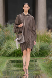 Runway model in a loose brown hooded dress with white drawstrings, carrying a taupe shoulder bag and wearing black strappy sandals.
