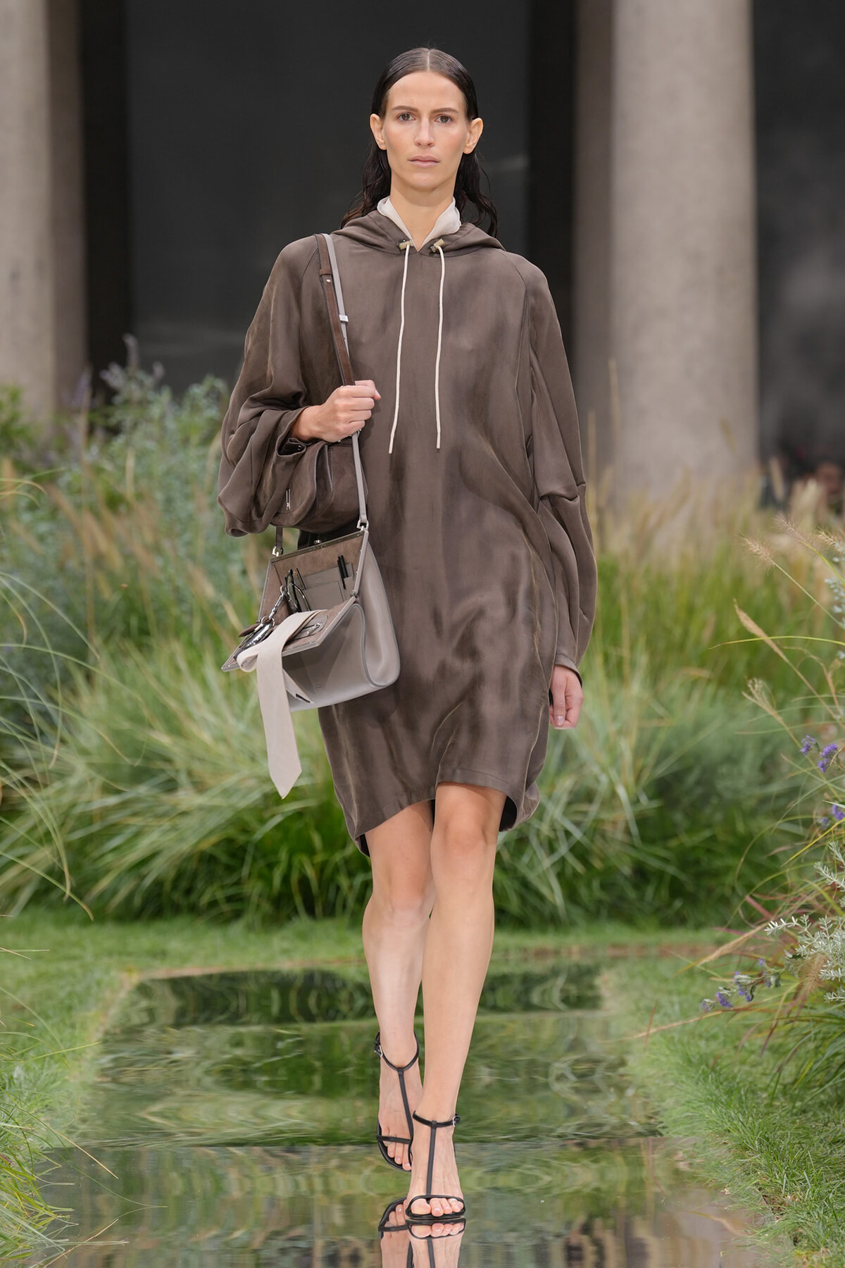 Runway model in a loose brown hooded dress with white drawstrings, carrying a taupe shoulder bag and wearing black strappy sandals.
