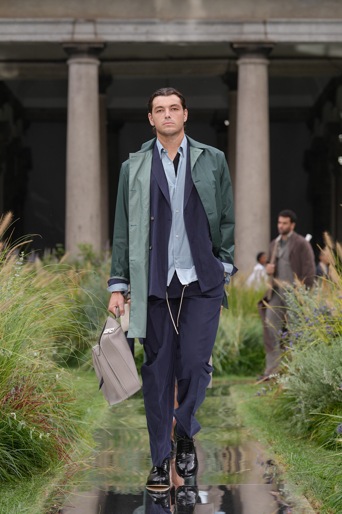 Male model walking a mirrored runway wearing a green trench over navy and light blue layered suit, carrying a gray briefcase.