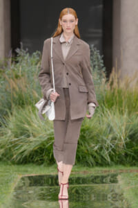Model in a taupe oversized blazer and matching cropped trousers with a beige blouse, carrying a white shoulder bag on an outdoor runway.Set in a garden-like backdrop with tall grass.