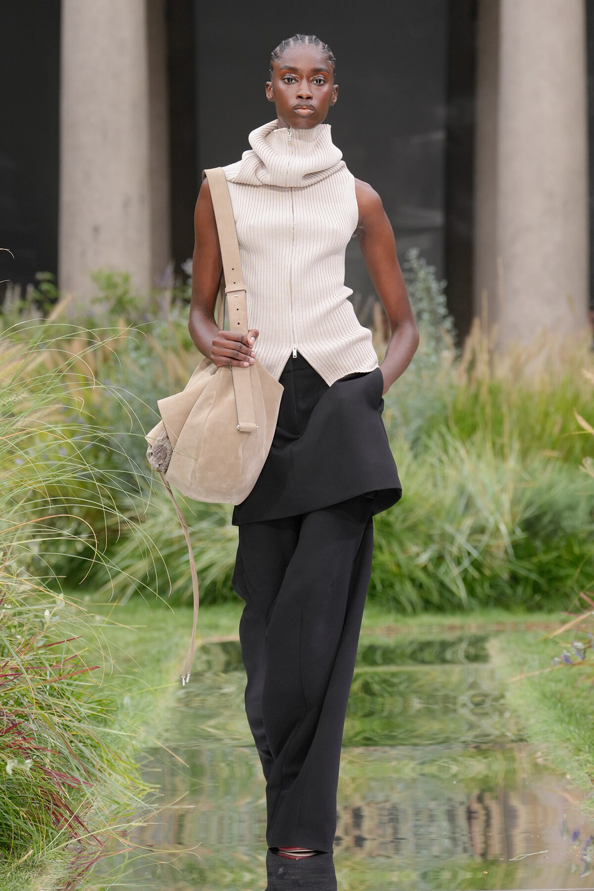 Model walking outdoors in a beige sleeveless knit turtleneck and black layered pants, carrying a tan shoulder bag.