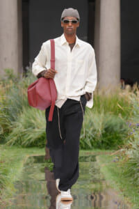 Male model in a white oversized shirt, black pants, gray cap, white sunglasses, and a red shoulder bag walking a reflective runway.
