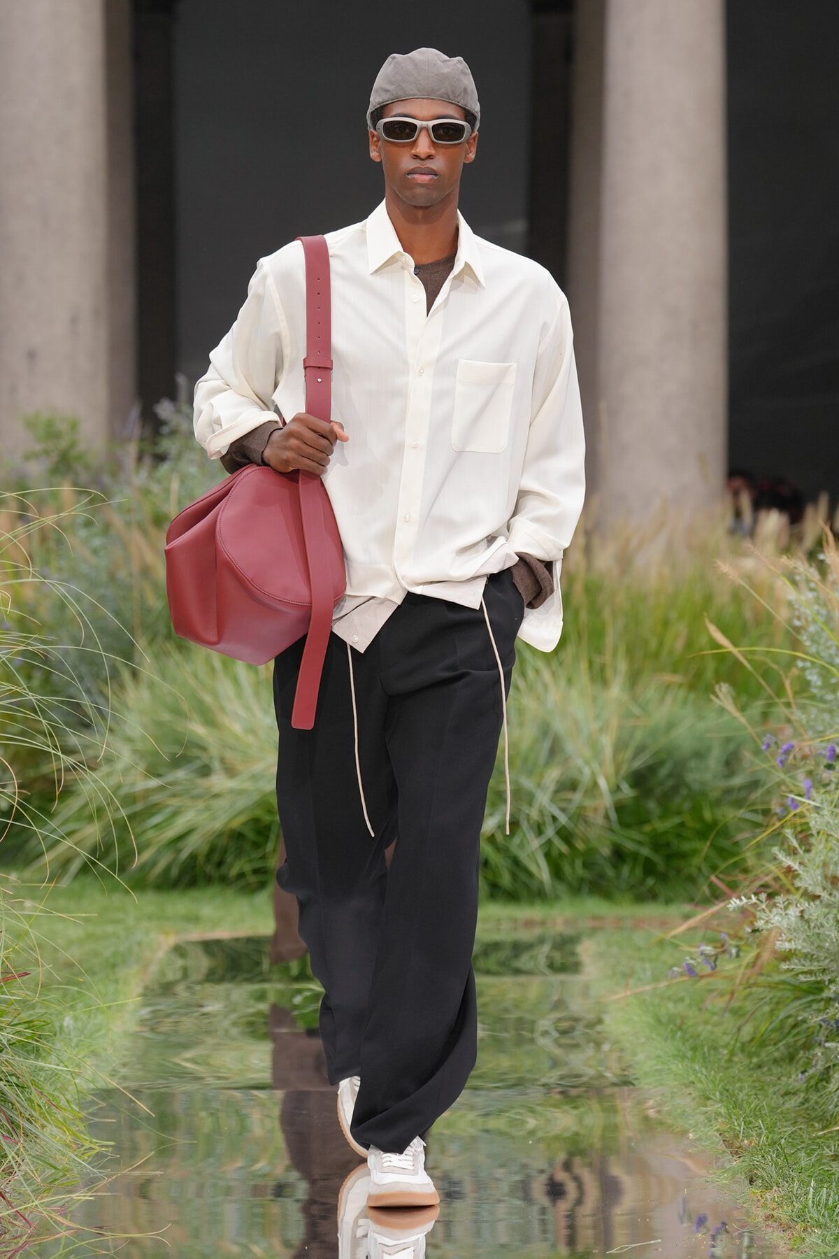 Male model in a white oversized shirt, black pants, gray cap, white sunglasses, and a red shoulder bag walking a reflective runway.