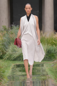 Female model walking on a runway in a flowing light-gray draped dress with a black neckline, holding a red clutch outdoors.