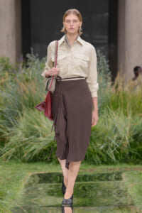 Female model walking outdoors on a fashion show runway, wearing a beige button-down shirt tucked into a brown midi wrap skirt with a thin belt, carrying a red handbag over the shoulder and wearing black mules, with greenery in the background.