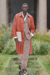 Male model walking outdoors in an orange leather overcoat over a tan suit, carrying a beige portfolio under his arm.