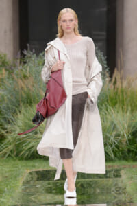 Model walking on a runway wearing a long beige trench, ribbed beige sweater, brown leather pants, white heels, and a burgundy oversized bag.