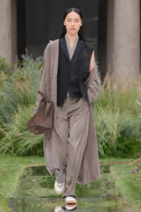 Female model walking outdoors in a taupe long coat over a black vest and matching pants, carrying a brown handbag on her left arm.