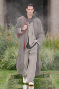 Male model in a layered gray coat and light suit walking through a misty outdoor garden with a red shoulder bag.