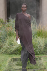 Model walking outdoors in a one-shoulder, dark plum gown with a white clutch, beside a reflective water feature.