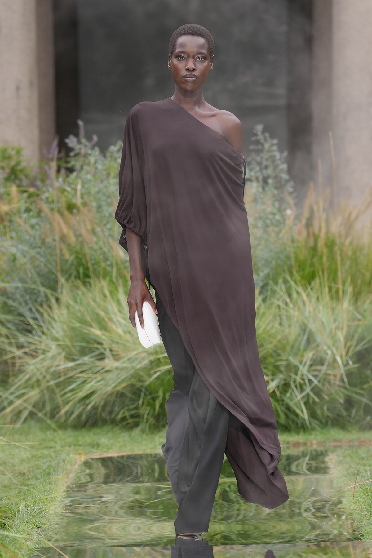 Model walking outdoors in a one-shoulder, dark plum gown with a white clutch, beside a reflective water feature.