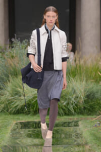 Model walking a fashion runway outdoors in a white jacket, black top, gray skirt, taupe tights and beige shoes, carrying a navy bag.