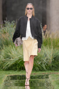 Model walking outdoors in a black bomber jacket, white blouse, beige knee-length skirt, strappy sandals, and sunglasses carrying a taupe clutch.