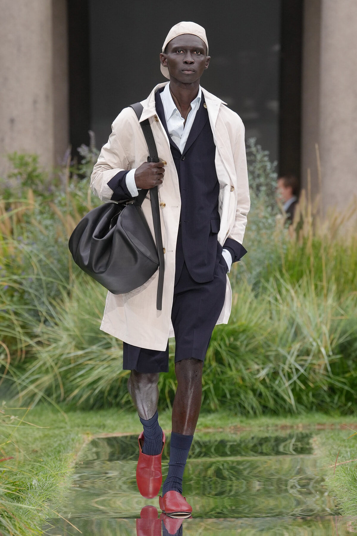 Male model walking outdoors in a light trench coat over a navy suit, carrying a large black bag.