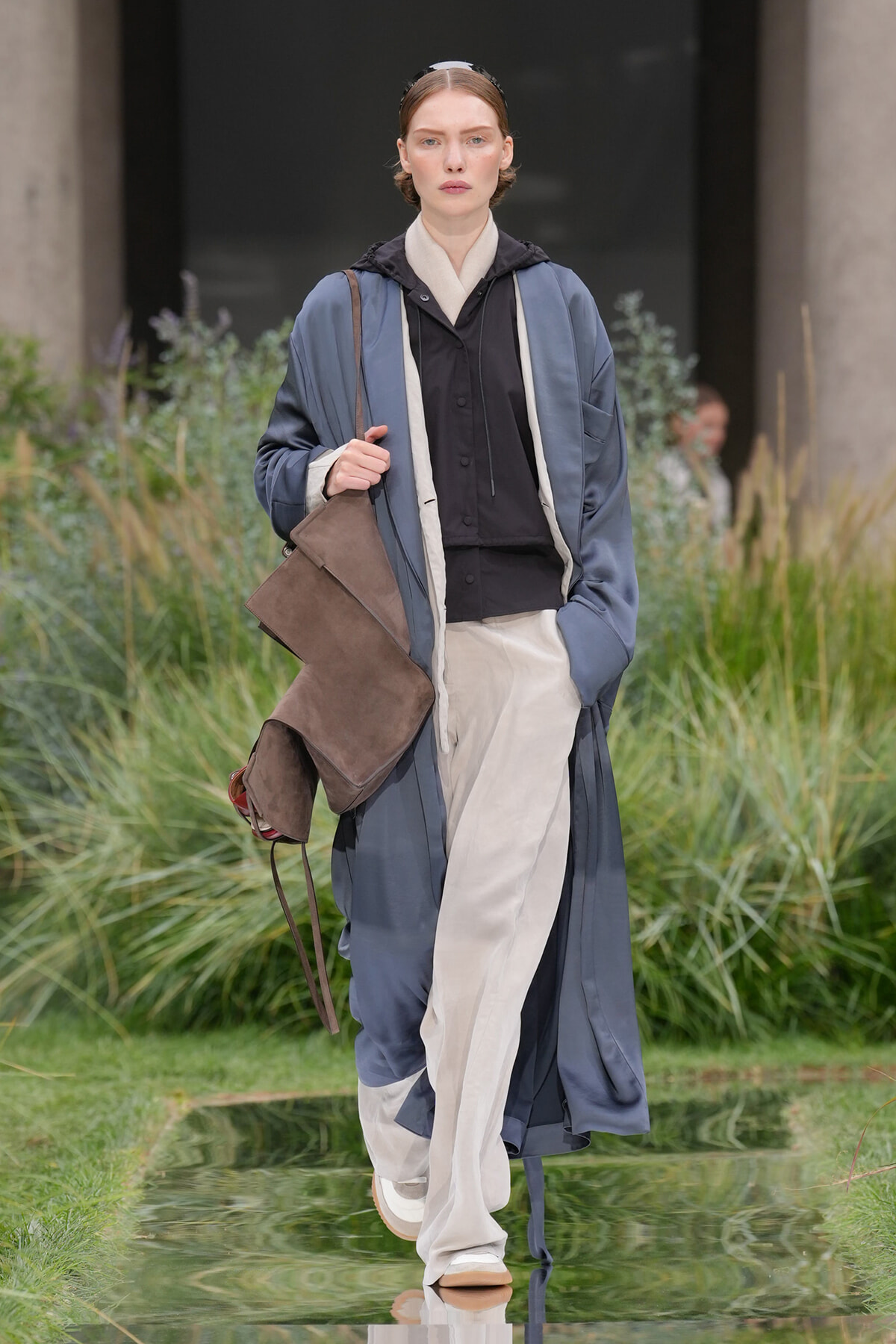 Model walking an outdoor runway in a blue oversized coat over a black top and beige wide-leg trousers, carrying a brown suede bag.