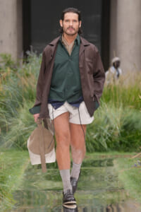 Man walking through a grassy, water-filled path in a brown coat over a green shirt, beige shorts, gray socks, and black loafers, carrying a beige bag.