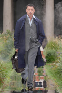Man in gray suit and navy overcoat walks on a reflective runway, carrying a dark leather briefcase.