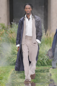 Male model in a navy trench over a beige scarf, striped shirt, and beige pants walking on an outdoor runway.