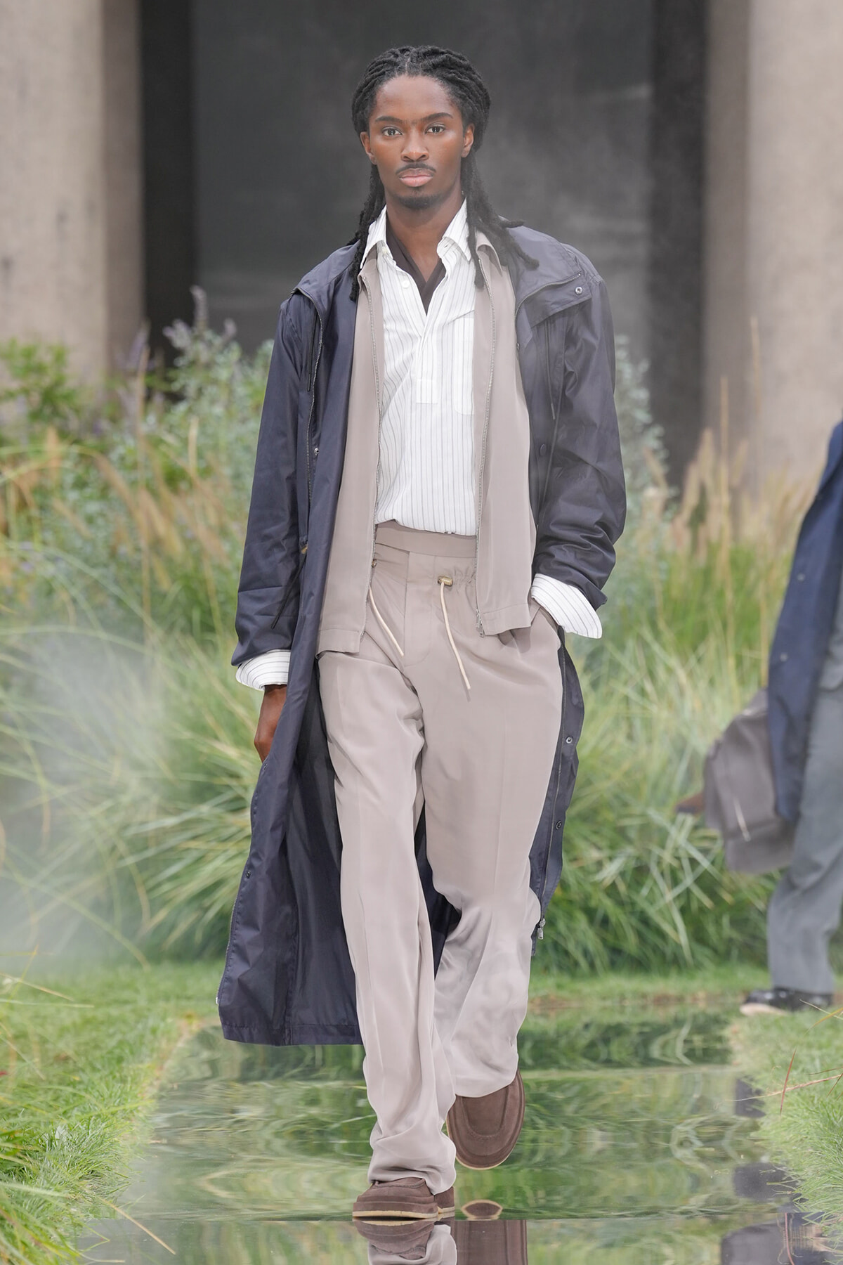 Male model in a navy trench over a beige scarf, striped shirt, and beige pants walking on an outdoor runway.
