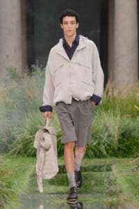 Male model walking along a reflective runway outdoors in a loose light beige jacket, dark shirt, gray shorts, and carrying a beige duffel bag.