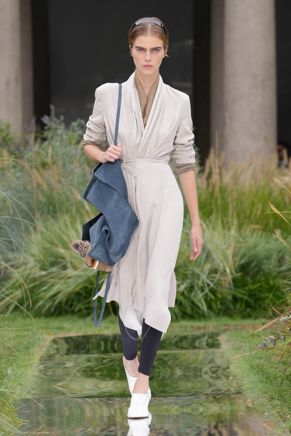 Model walking outdoors in a beige draped trench dress with a blue crossbody bag and white mules, surrounded by greenery.