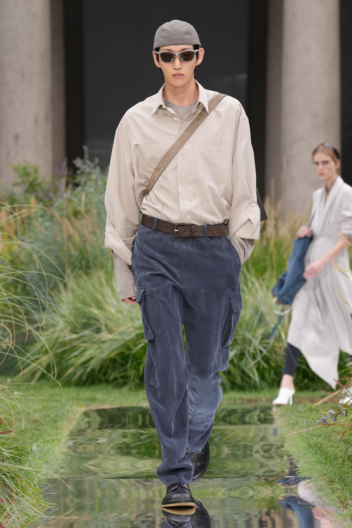 Male runway model in a beige shirt, blue cargo pants, crossbody bag, cap and sunglasses walking on a reflective outdoor catwalk.
