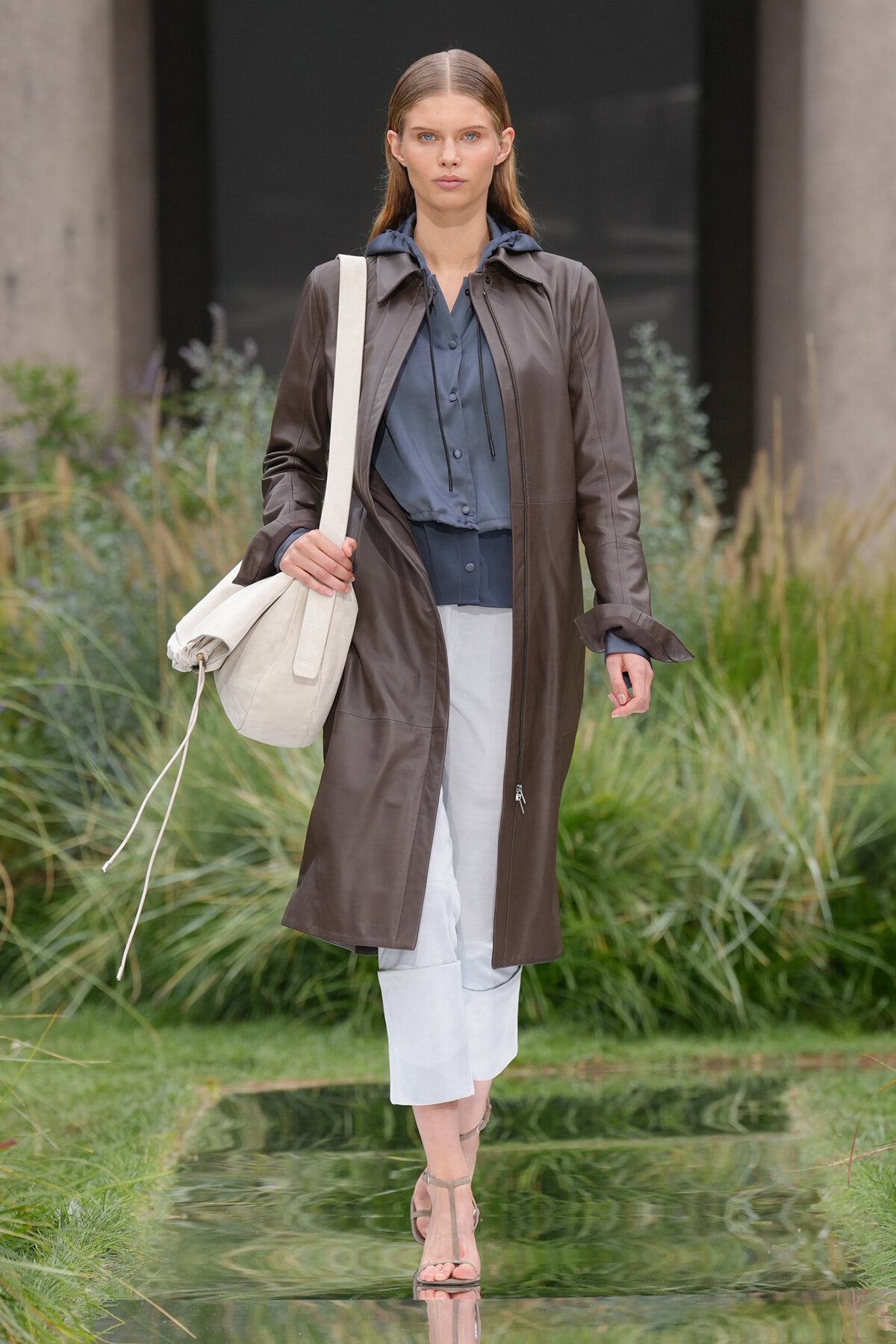 Model walking outdoors on a fashion runway wearing a knee-length brown leather coat over a blue blouse, white cropped pants, and a beige tote bag.