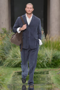 Man in a navy blue suit with a white shirt walks along a reflective water feature carrying a brown shoulder bag in a garden setting.