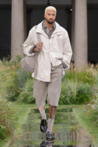 Male model with bleached blond hair walks a runway in a light beige oversized jacket, gray shirt, and matching shorts, carrying a large gray bag.