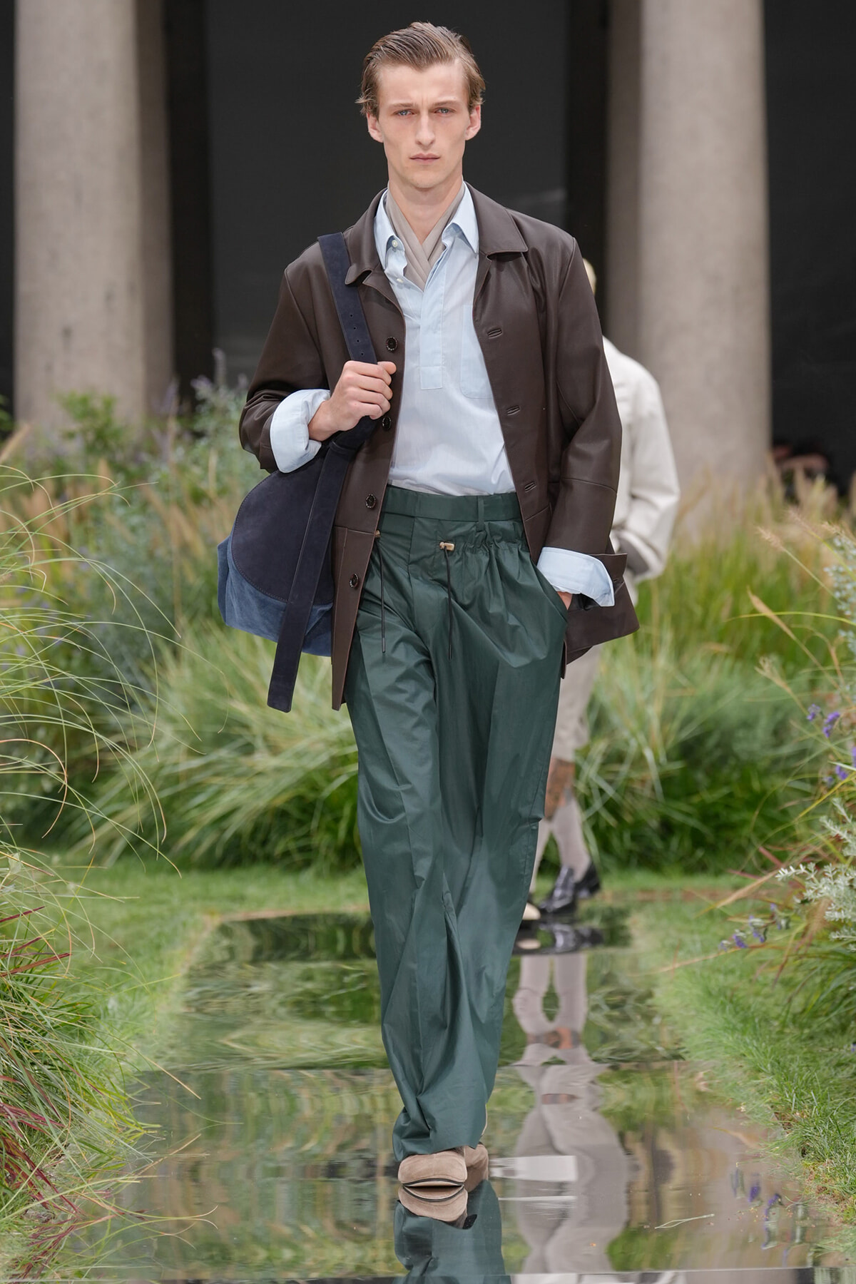 Male model walking along an outdoor runway with a navy bag, wearing a brown leather jacket, light blue shirt, and dark green drawstring trousers.