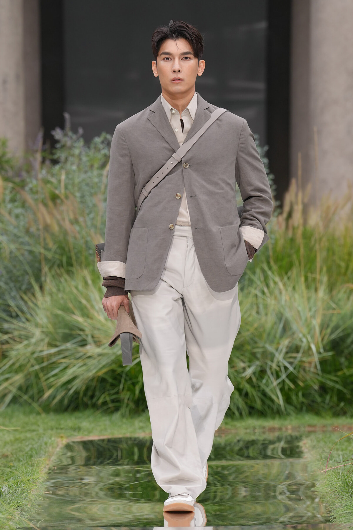 Male model walking outdoors in a gray blazer and light trousers, with a crossbody bag and hat in hand.