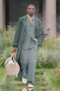 Male model walking a fashion show in a green layered suit (coat, vest, trousers) holding a beige duffel bag on a reflective garden runway with plants in the background.