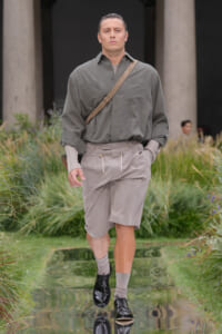 Male model struts down a garden runway in a muted gray shirt, beige shorts, and a tan crossbody strap, with black shoes and gray socks.