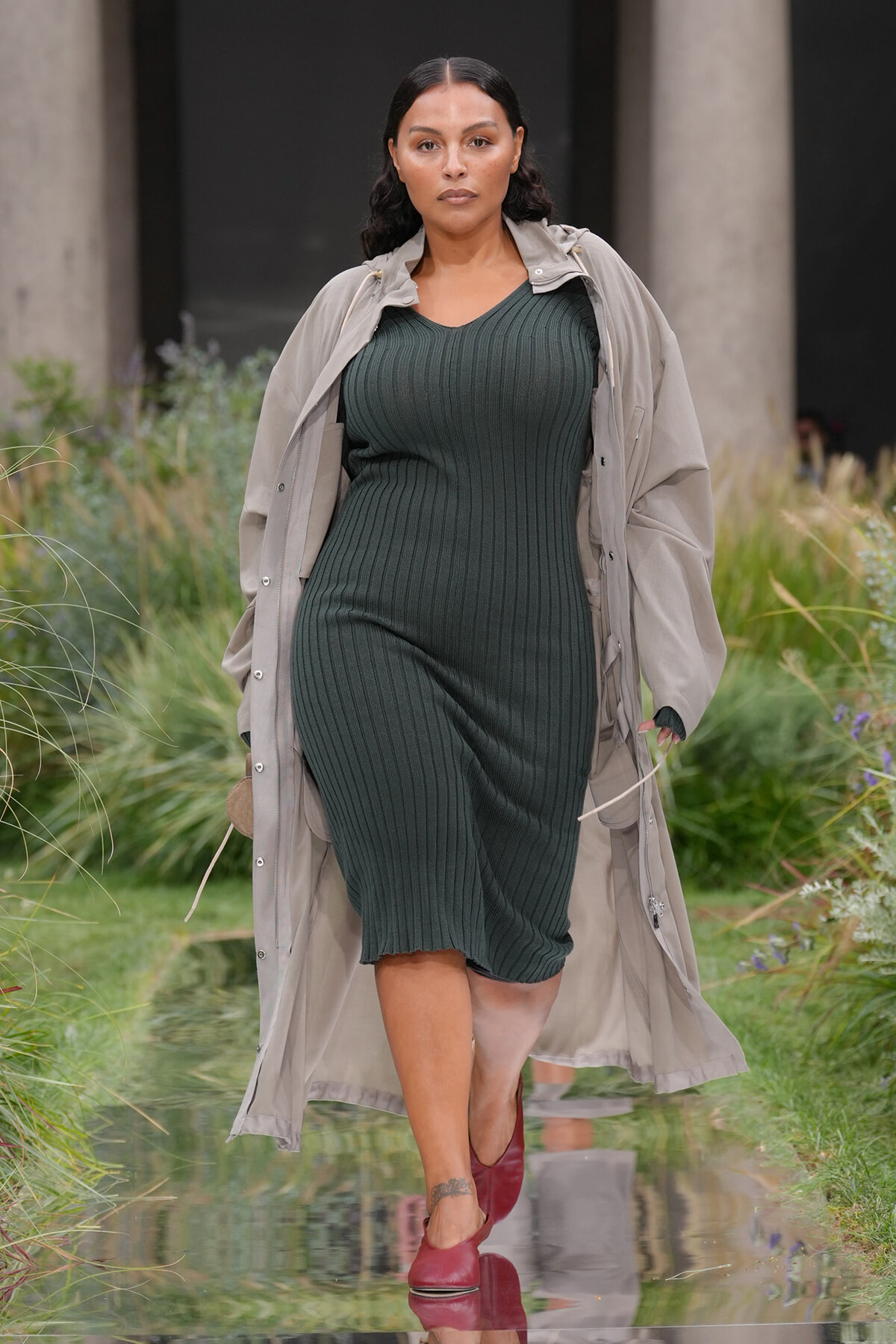 Model walking on a runway outdoors in a dark green ribbed midi dress and beige trench coat, red shoes, serious expression.