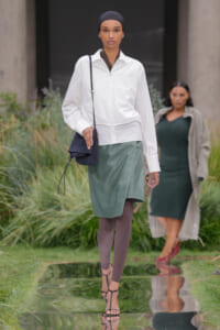 Model walking on a reflective runway wearing a white zip-front jacket, a muted green wrap skirt, taupe tights, and strappy sandals, carrying a small navy crossbody bag and wearing a black head wrap.