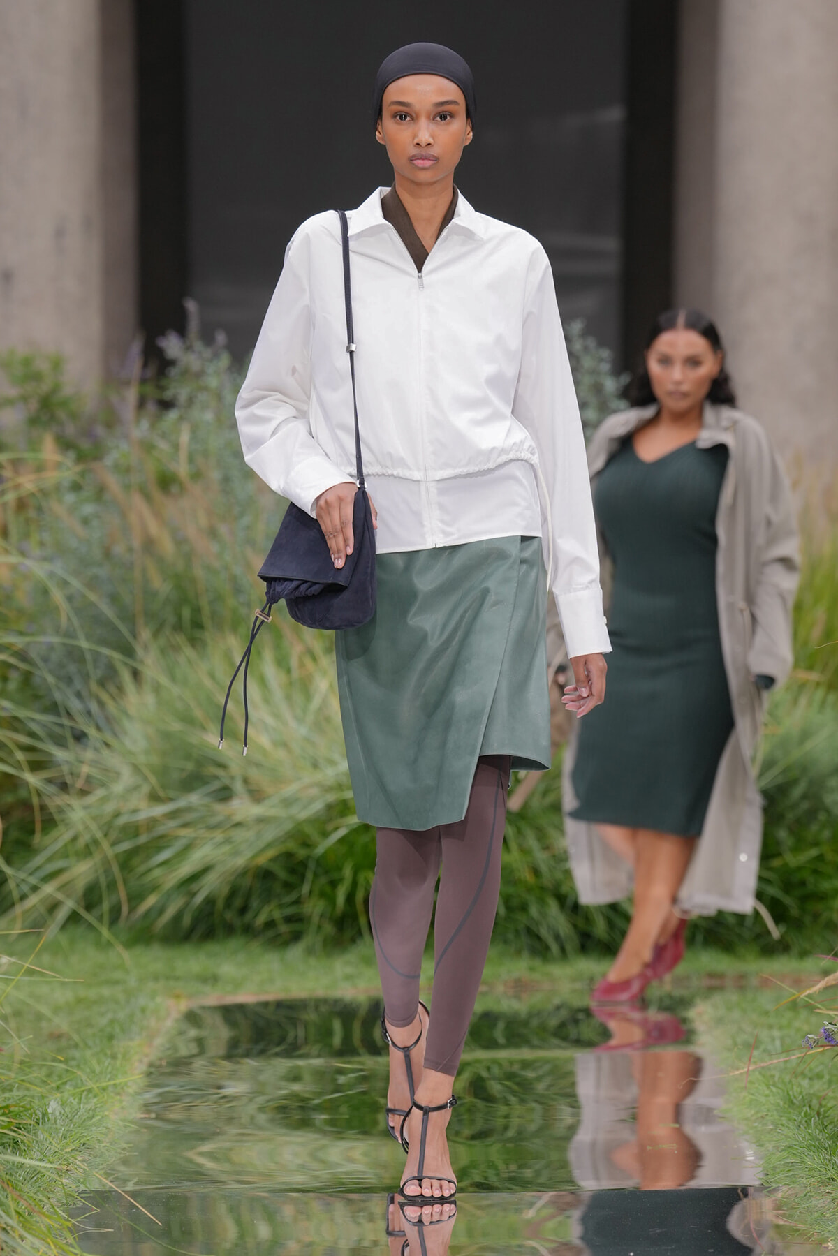 Model walking on a reflective runway wearing a white zip-front jacket, a muted green wrap skirt, taupe tights, and strappy sandals, carrying a small navy crossbody bag and wearing a black head wrap.