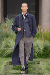 Male model walking a runway in a navy long coat over a gray jacket and beige pants, carrying a green shoulder bag.
