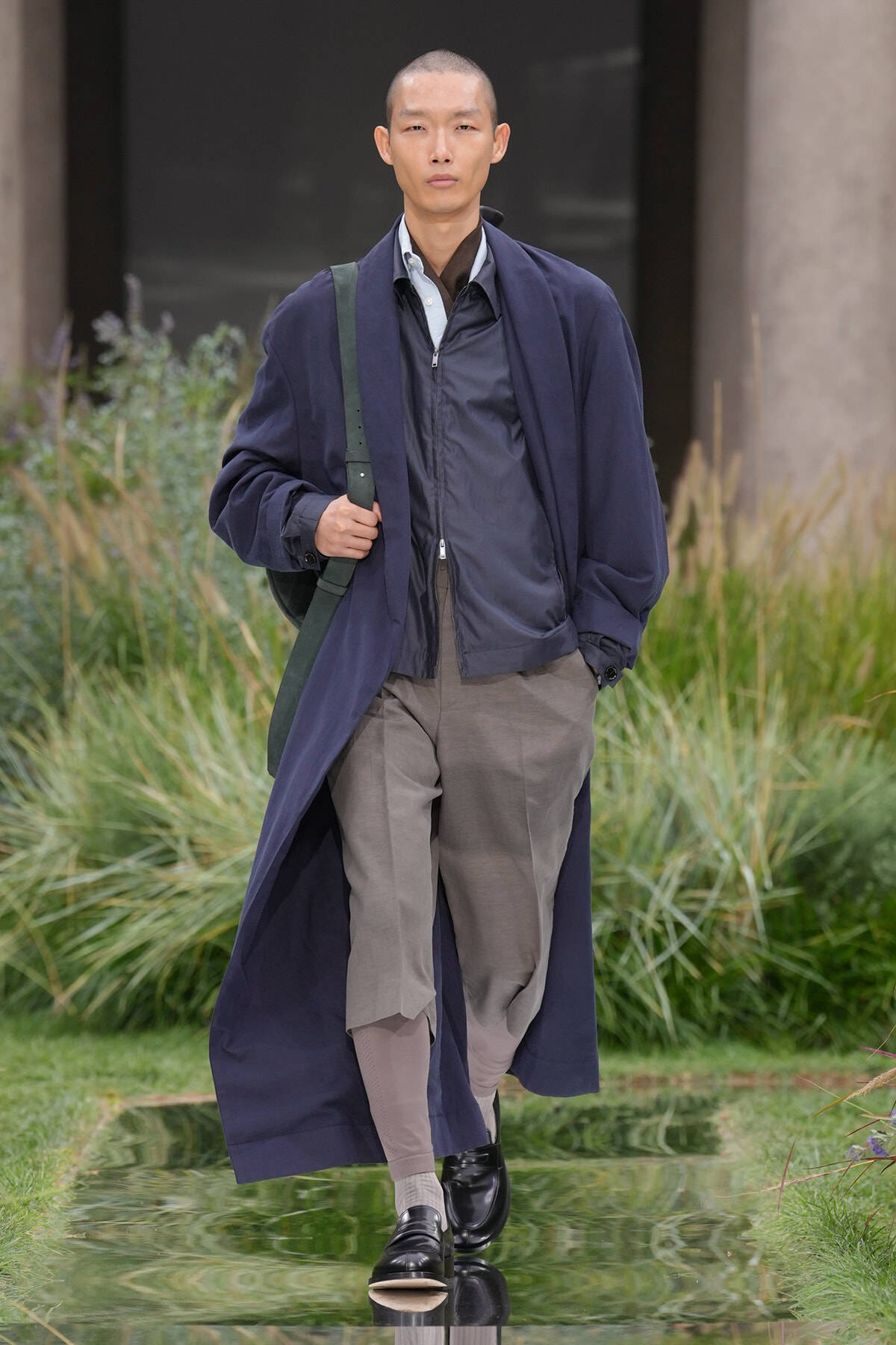 Male model walking a runway in a navy long coat over a gray jacket and beige pants, carrying a green shoulder bag.