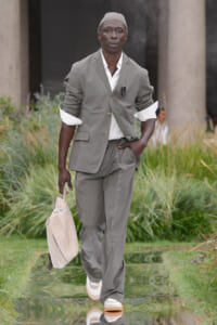 Male model in a gray suit walks along a reflective outdoor path, carrying a light beige briefcase and wearing a matching cap.
