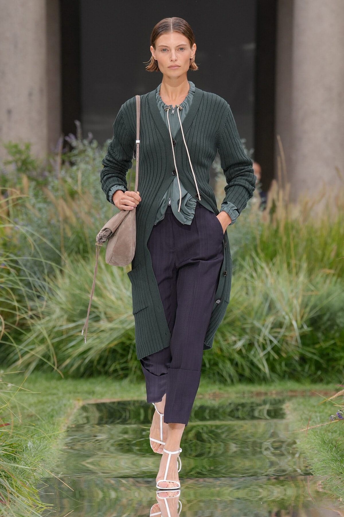 Model walking outdoors in a dark green ribbed cardigan over a teal blouse, navy cropped trousers, white strappy sandals, and a beige shoulder bag.