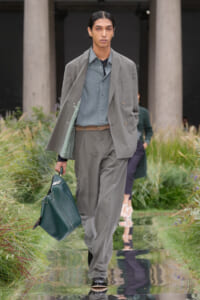 Male model walking a runway in a light gray suit with a blue shirt, carrying a dark green briefcase-style bag.