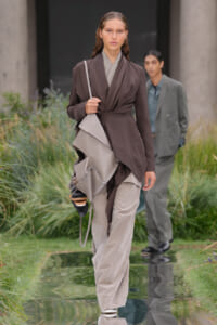 Model walks down an outdoor runway wearing a dark brown, wrapped-front coat with a light taupe draped layer and loose beige pants; she carries a light gray angular shoulder bag. A second model in a gray suit stands in the background near tall plants.