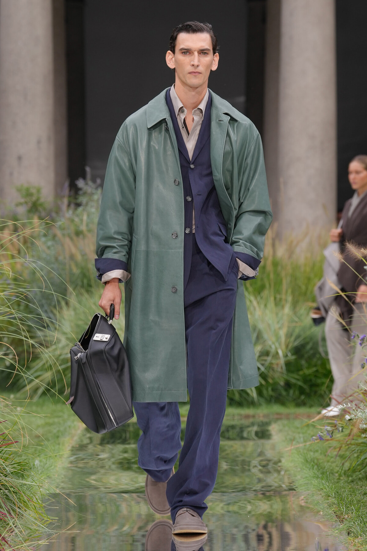 Male model walks a fashion runway wearing a green oversized coat over a navy suit, and holds a black briefcase.
