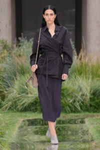 Model walking outdoors on a runway in a dark wrap blouse, matching wide-leg pants, beige crossbody bag, and gray mules, with a calm, focused expression.